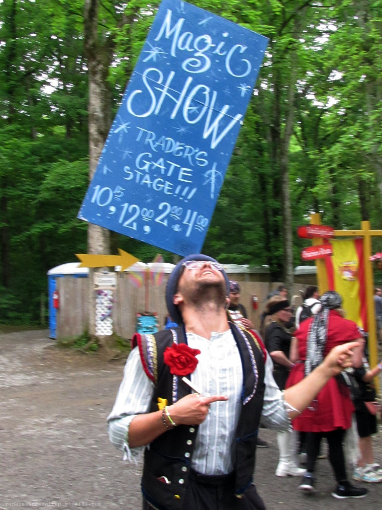 Man balancing aa large sign on his forehead. The sign says Magic, he's pointing to the stage  on which he's about to do a show.