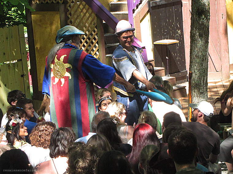 Two mens standing on benches in audience for a show. They are pointing bowling pin at a man they want to come on stage.
