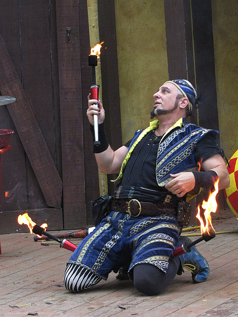 Man intights. Crouching on a stage. he is holding a flaming torch. with more flaming torches held beding his knees.