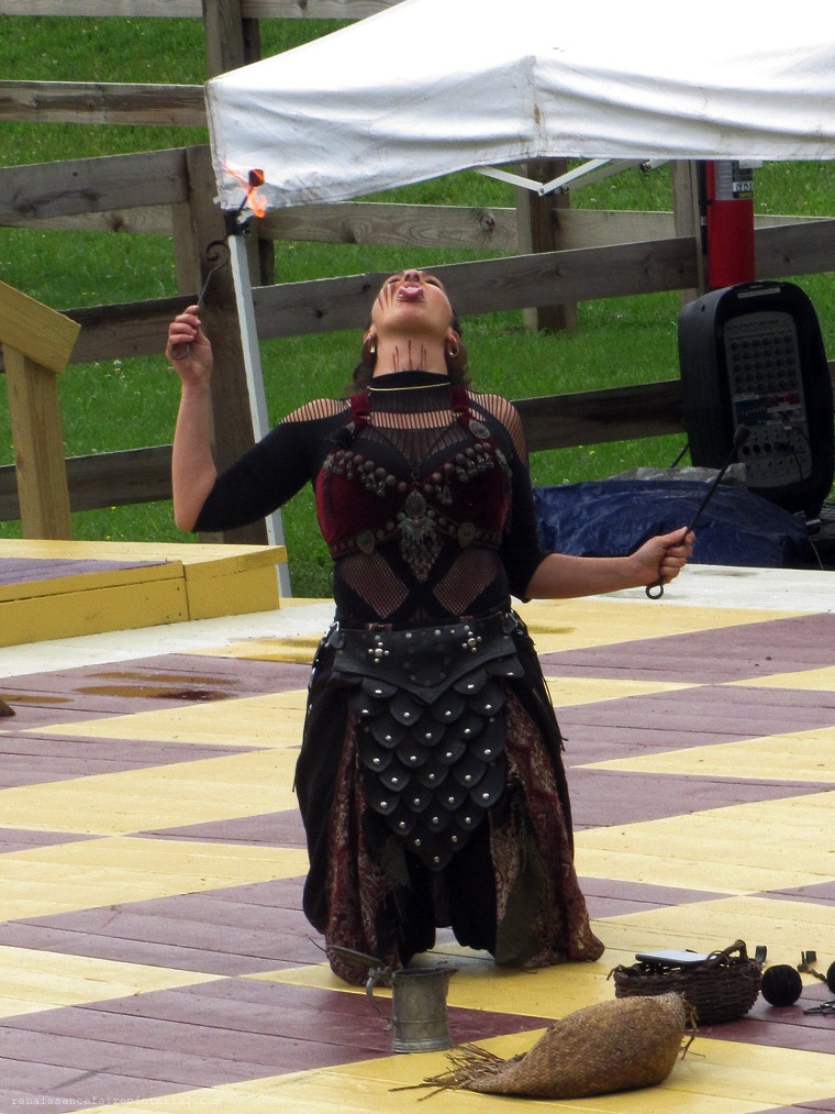 Woman kneeling with her head thrown back. she's holding two sticks one with a flame on it one without. her tounge has a flame on it. she just tranfered the flmae fromstick to her tounge.