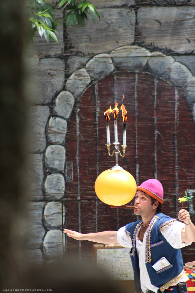 man with a stick in his mouth. on the stick he is balacing an ballon on top of the ballon in a canslerbra with three lit candles. he' about to pop the ballon,
